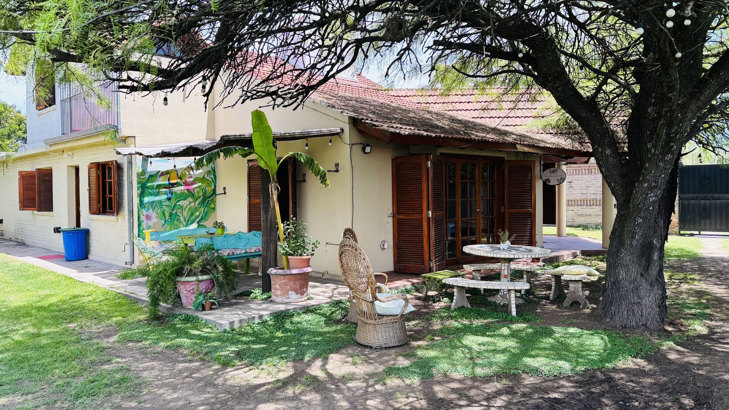 Jardín con un mural, un banano y mesa bajo el algarrobo